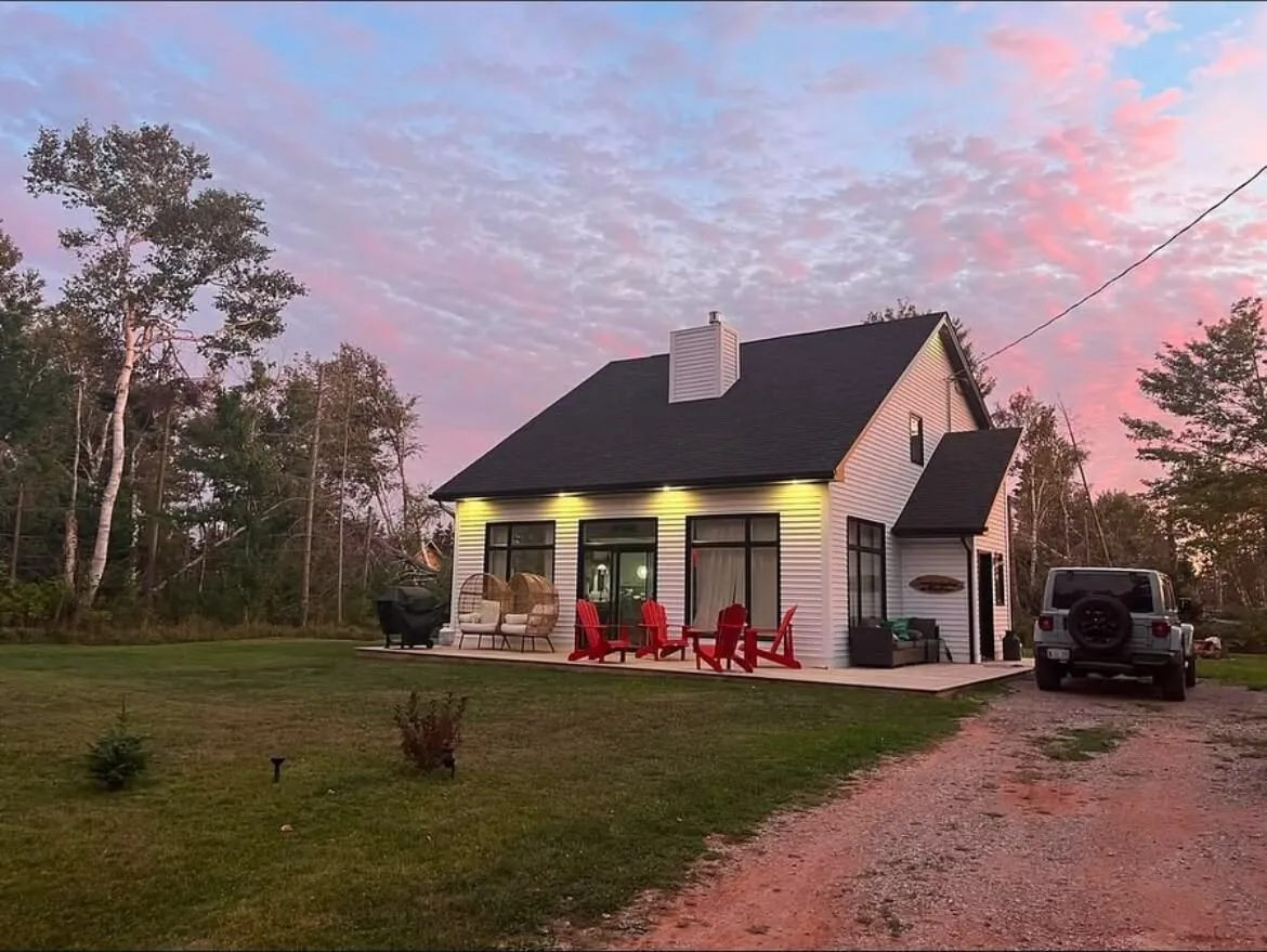 The cottage at dusk under a pink sky, soffit lights on and red chairs on the front deck